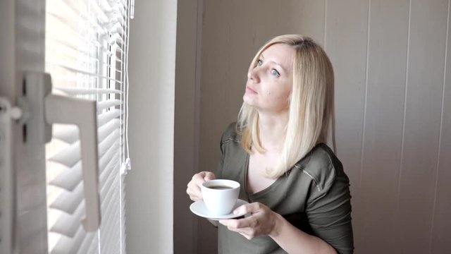 Young sad thinkful beautiful blond woman standing near the window with blinds in the morning and drinking coffee
