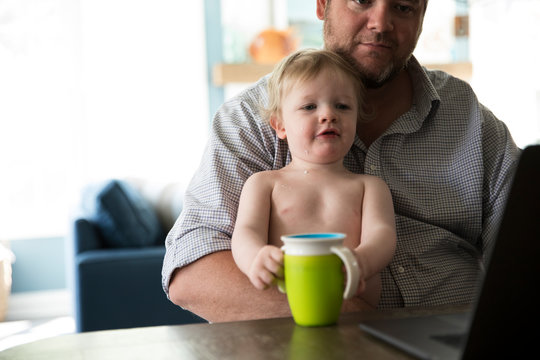 Dad Working on Computer, Holding Toddler Boy Drinking From Sippy Cup