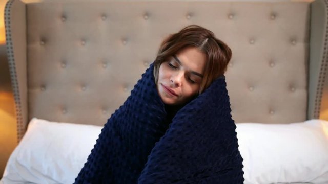 Woman wrapped in a blue blanket. Cosy sitting on the bed and smiling.