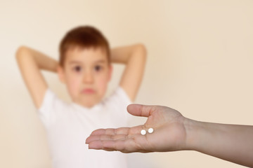Naklejka premium female hand stretches two white pills on the palm on a blurred background of a boy who raised his hands behind his head with a distressed face, light background
