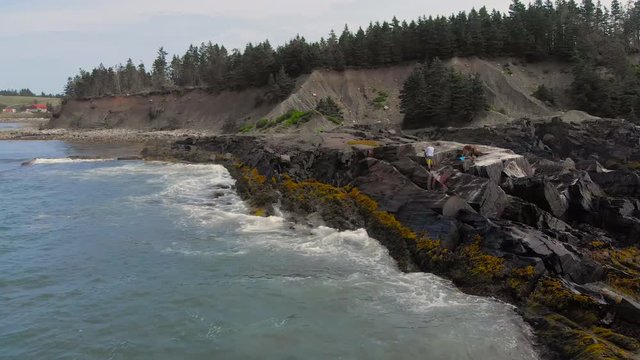 Cinematic drone / aerial footage moving back showing a bay, someone taking pictures on the rocks and a dog in the cost of Kingsburg, Nova Scotia, Canada during summer season.