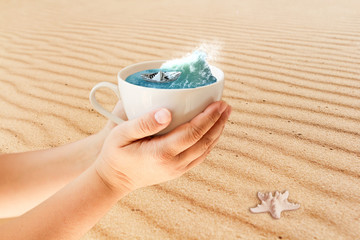 Paper boat from a newspaper in a white mug in hands, where the blue sea is splashing against the background of sand dunes