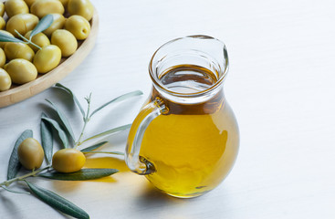 Glass bottle of olive oil and olive tree branch, raw turkish green olive seeds and leaves on white table. green olives background, olivae oleum
