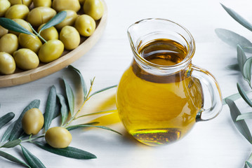 Glass bottle of olive oil and olive tree branch, raw turkish green olive seeds and leaves on white table. green olives background, olivae oleum