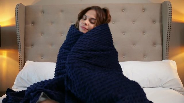 Woman wrapped in a blue blanket. Cosy sitting on the bed and smiling.