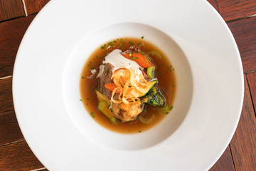 White plate with beef soup. Pieces of beef meat with dumplings, carrot, slices of celery, cream, embellished with parsley, chives and fried onion strips. Top view.