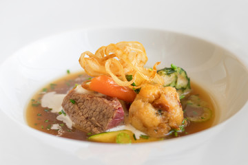 White plate with beef soup. Pieces of beef meat with dumplings, carrot, slices of celery, cream, embellished with parsley, chives and fried onion strips. Closeup.