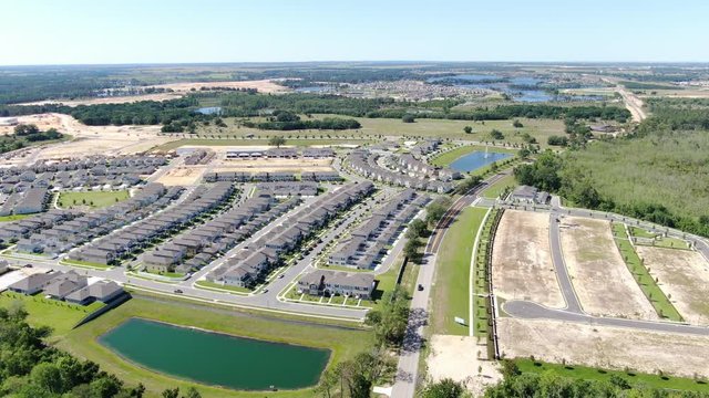 Flying over newly constructed neighborhood surrounded by green woods and rolling fields on a summer afternoon