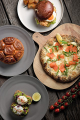 Top down lunch table with a salmon pizza, a cinnamon roll, a chicken burger and some poached eggs