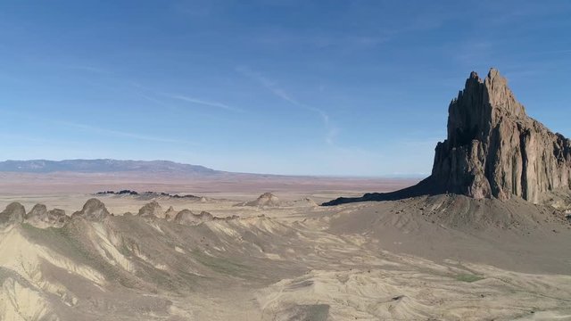 A slow aerial flyby of a towering rocky cliff face. Depicted through a steady wide shot that drifts in a rightwards direction to reveal the vast expanse of the desert surroundings below.