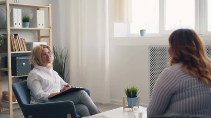 Successful psychologist mature lady is talking to overweight young woman during therapy session in modern clinic. People, conversation and mental health concept.