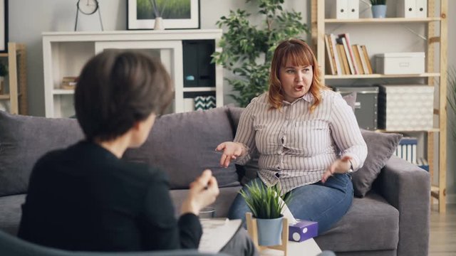 Unhappy obese woman in casual clothing shirt and jeans is telling psychologist about personal problems during session then taking paper tissue touching nose.