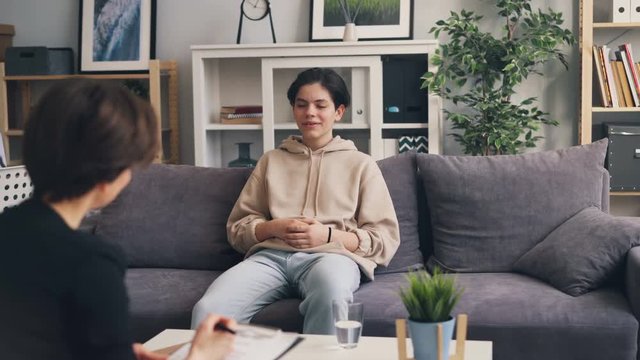 Emotional teenager dark-haired boy is smiling talking to psychotherapist during session in clinic listening to specialist's advice. Youth and psychology concept.