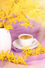 A cup of coffee stands on a textural silk napkin of a delicate color by a vase with flowering twigs  of forsythia, the sun's rays fall on the table