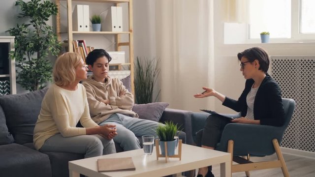 Troubled teenager is visiting therapist with mother, worried mature woman is talking asking advice while boy is sitting with arms crossed. Family problems and psychology concept.