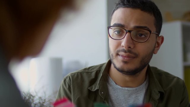 Over-shoulder close-up shot of young Arabic graphic designer wearing glasses, who is demonstrating colour cards to colleague and trying to persuade them
