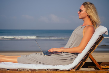 businesswoman working on a laptop while sitting in a lounger by the sea on a white sandy beach. freelancing remote work workaholism concept.