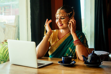 indian beautiful businesswoman in green sari and tika working on a modern laptop in the cafe...
