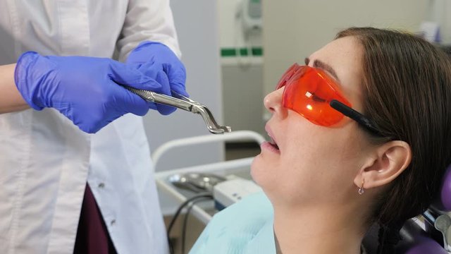 Portrait of young woman afraid to remove a tooth sitting in dental chair while the doctor stands next to the patient, holding forceps in hands.
