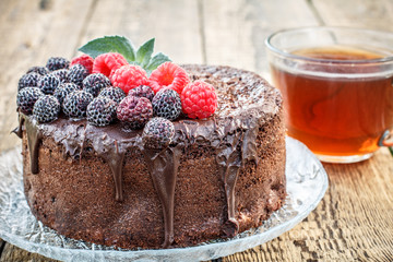 Homemade chocolate cake decorated with black and red raspberries on glass plate with cup of tea