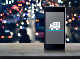 Free delivery truck flat icon on modern smart mobile phone screen on wooden table over blur colorful night light traffic jam road with cars in city, Business transportation online concept