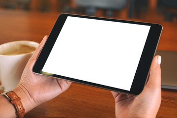 Mockup image of hands holding black tablet pc with blank white screen with laptop and coffee cup on wooden table
