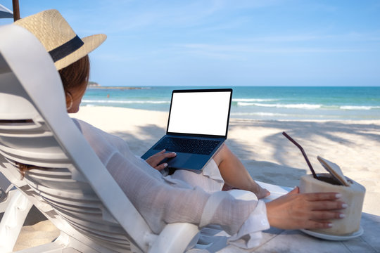 Mockup image of a woman holding and using laptop computer with blank desktop screen while laying down on beach chair and drinking coconut juice on the beach
