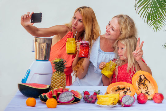 Family In The Kitchen During Breakfast At Home.three Blonde Sisters Cooking Smoothies In A Blender.tropical Summer Theme. Mom And Daughter Taking Pictures Selfie Portrait On Camera Smartphone