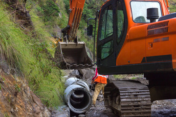 big orange excavator lifts a concrete pipe on a mountain road