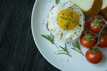 Beef steak with fried egg in spices. Decorated with rosemary, fresh cherry and slices of bread. Filed on a white plate. Dark wooden background. View from above.