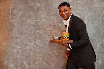Respectable young african american man in black suit hold tray with double burger against gray wall.
