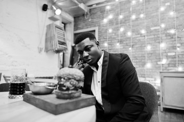  Respectable young african american man in black suit sitting in restaurant with tasty double burger and soda drink. Speaking at phone before eat.