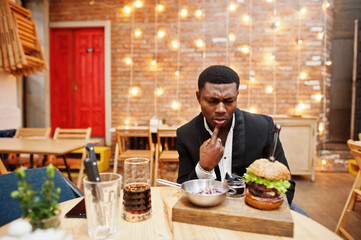 Eat or not? Respectable young african american man in black suit sitting in restaurant with tasty double burger and soda drink.