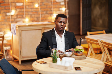Respectable young african american man in black suit sitting in restaurant with tasty double burger and soda drink.
