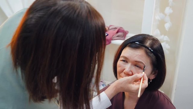 Makeup artist doing makeup eyebrows with cosmetic brush to elderly woman. Visagiste shaping and coloring eyebrows of mature woman in beauty studio. Beauty and makeup concept