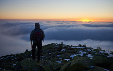 Fototapeta premium Back view of tourist in warm jacket with backpack and trekking sticks on rocky mountain peak on background of valley covered with white clouds stretching to horizon and bright morning sky at sunrise.