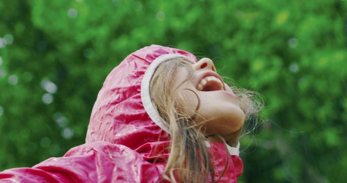Authentic shot of cute happy little girl in raincoat is having fun and enjoying the rain in a hot summer day in a green park.
