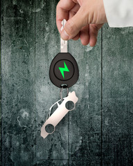 Green energy electric car and lightweight high strength speed concept. Businessman hand holding electric car key with sheet metal keyring in sports car shape, on dirty wall background.