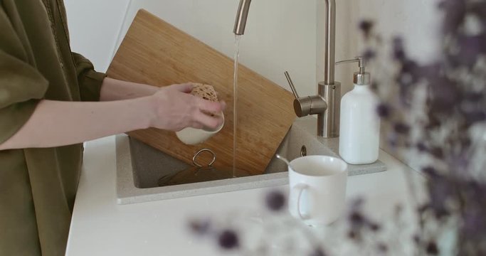 Woman washing dishes in the sink in the kitchen. In her hands she has a white mug, a spoon and a sponge with which she washes.