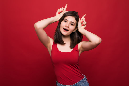 Portrait of funny young beautiful young woman standing with horns gesture hands on head and looking at camera isolated on red background.