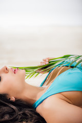 cropped view of sexy girl in bikini with eyes closed posing with green leaf while lying on beach with copy space