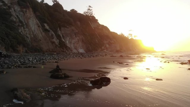 A man sitting in peaceful meditation and present awareness on a sandy beach with ocean waves at sunrise in Santa Barbara, California AERIAL DRONE.