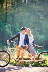Obraz premium Young happy couple tourists, bearded man and attractive woman close together at tandem double bicycle outdoors in summer park or forest on blurred sunny green trees dense foliage background.