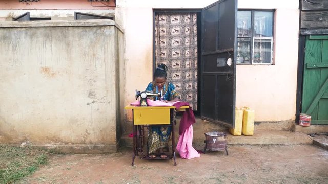 A wide shot of an African woman outside her small house sewing clothing on a manual tailoring machine.