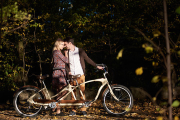 Young happy romantic couple, bearded man and attractive fashionable woman close together at tandem double bicycle outdoors in autumn park or forest on blurred dark trees dense foliage background.