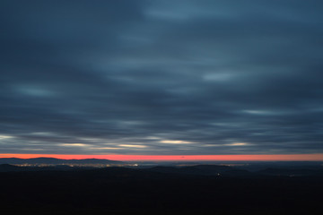 Night view of the lights of the city in the distance against the sky covered with clouds and a red strip of dawn on the horizon. High resolution and detail.