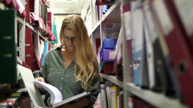 Blonde woman standing between shelves in archive searching binder for documents