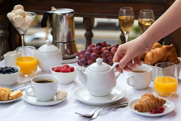 Woman hand holding teapot for making tea on hotel terrace. Luxury breakfast, coffee maker, teapot, cups, croissants, fruits, orange juice and champagne. Good morning concept background.