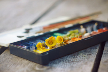 On the wooden table is an old vintage palette with cuvettes, a set of brushes and a few blossoming flowers coltsfoot