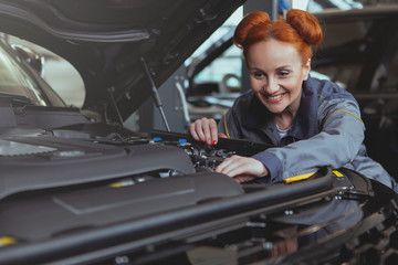 Fototapeta na wymiar Attractive cheerful red haired female mechanic smiling joyfully, repairing a car at the garage. Lovely woman automobile technician repairing car at the workshop, copy space